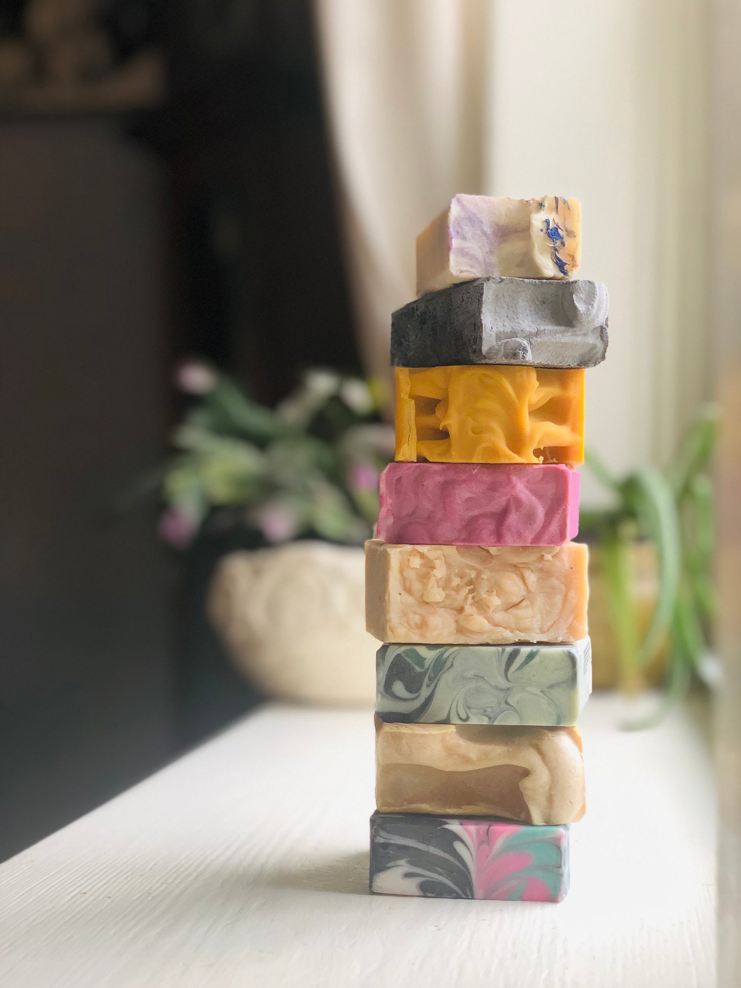 a stack of eight handmade soaps of a variety of colors and sizes sitting in a window sill, two plants in the background