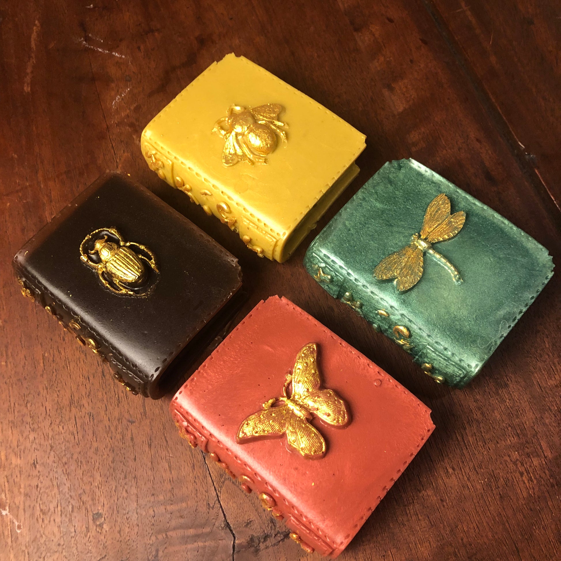 Four book shaped soaps with insects in gold on the cover. Insects are beetle on brown cover; bee on yellow cover; butterfly on orange cover and dragon fly on green cover. These are on a brown table. 